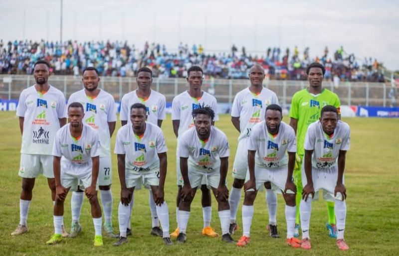 The Masters FC team in white kit lined up on the pitch with fans in the stand behind